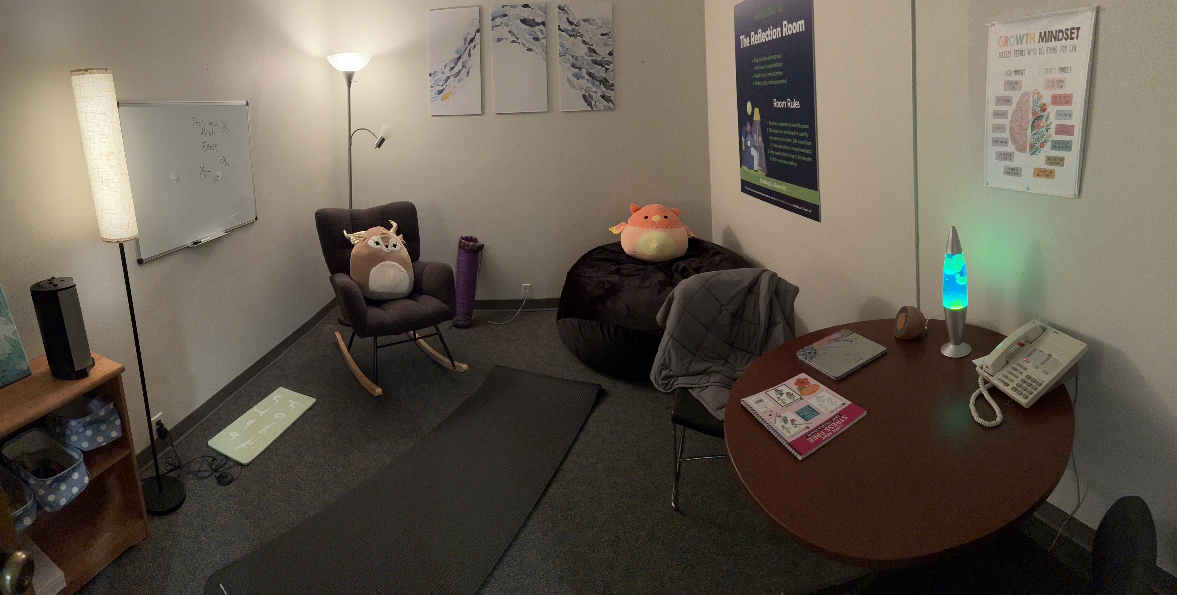 A wide shot of the reflection room, with soft lighting, yoga mats on the floor, a comfy rocking chair and beanbag with stuffed animals, a table with coloring books and lava lamp, and posters and artwork on the walls.